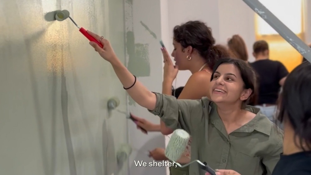A group of people painting a wall light green with rollers; one person in front is smiling. Subtitles read, "We shelter;".