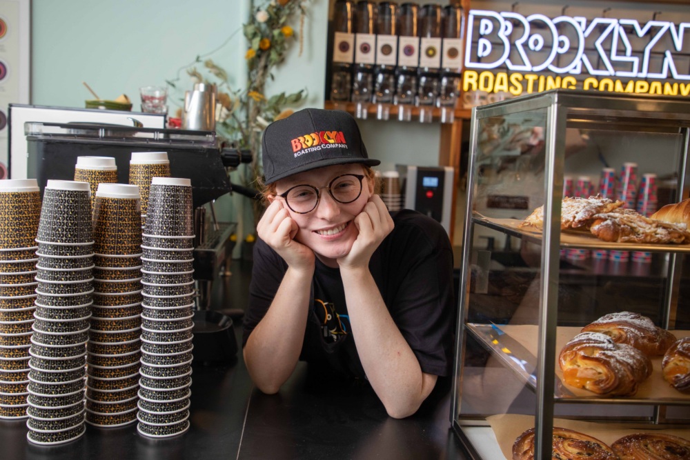 A person wearing glasses and a Brooklyn Roasting Company cap smiles behind a coffee counter with stacked cups and a pastry display case.