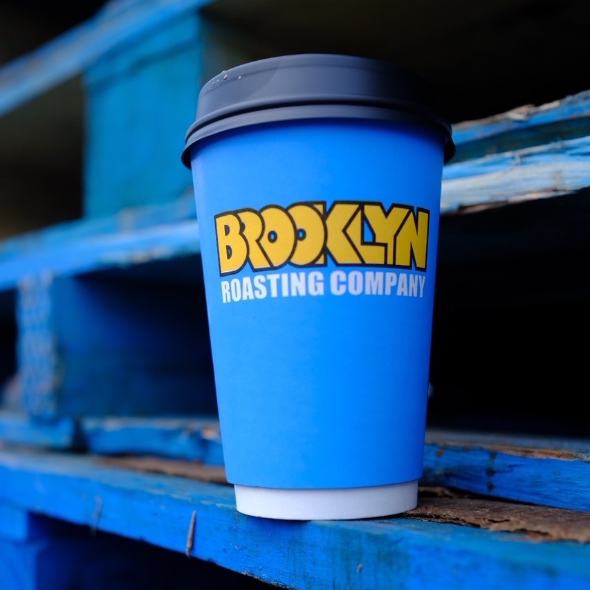 A blue Brooklyn Roasting Company coffee cup with a black lid sits on a stack of blue-painted wooden pallets.