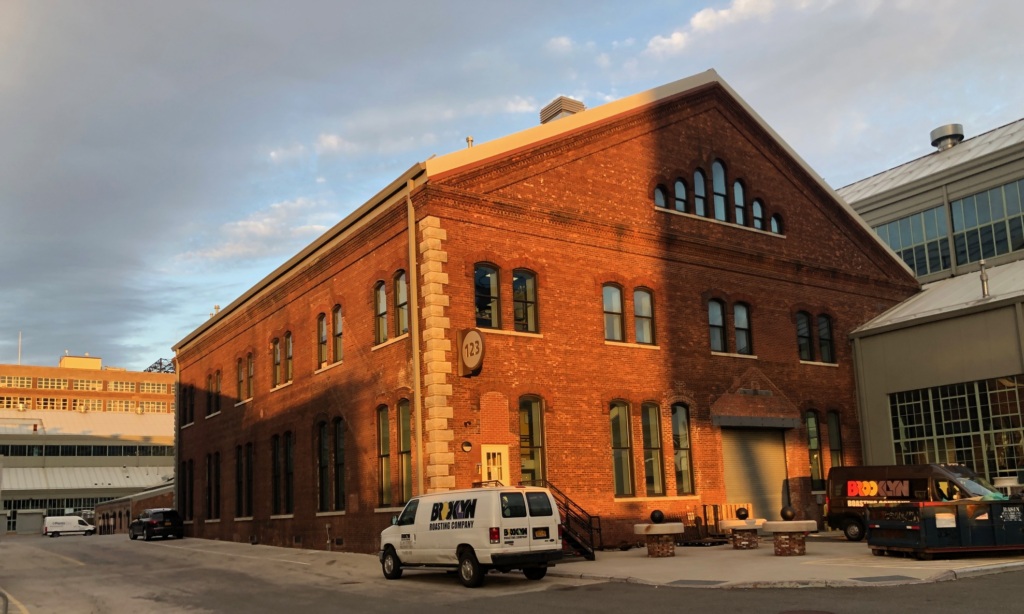 A two-story red brick industrial building with arched windows, a white van parked in front, and a large roll-up door, illuminated by late afternoon sunlight.