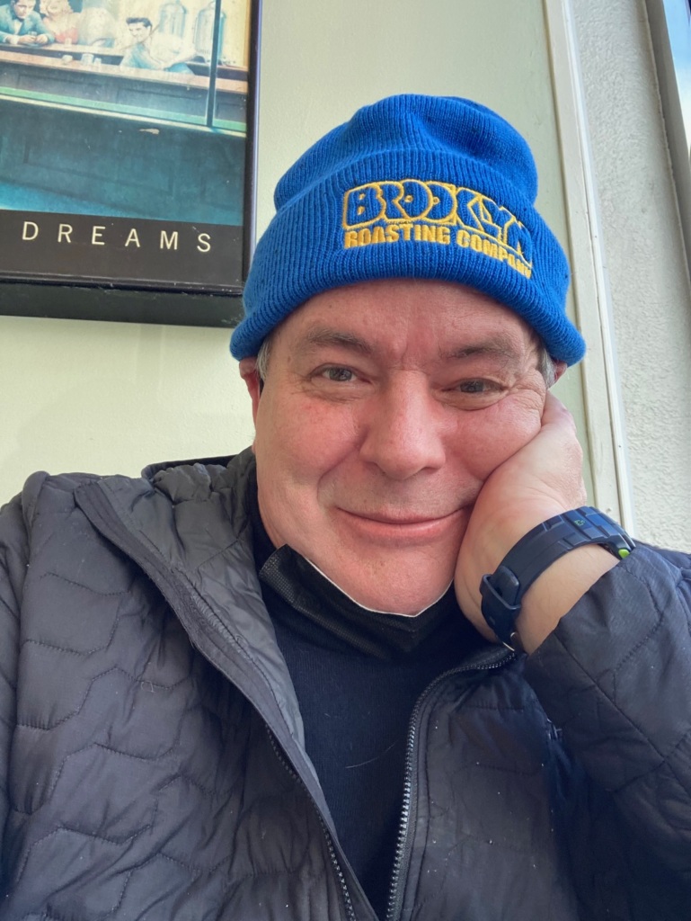 A man wearing a blue beanie and black jacket smiles at the camera, resting his head on his hand. A framed photo is visible on the wall behind him.