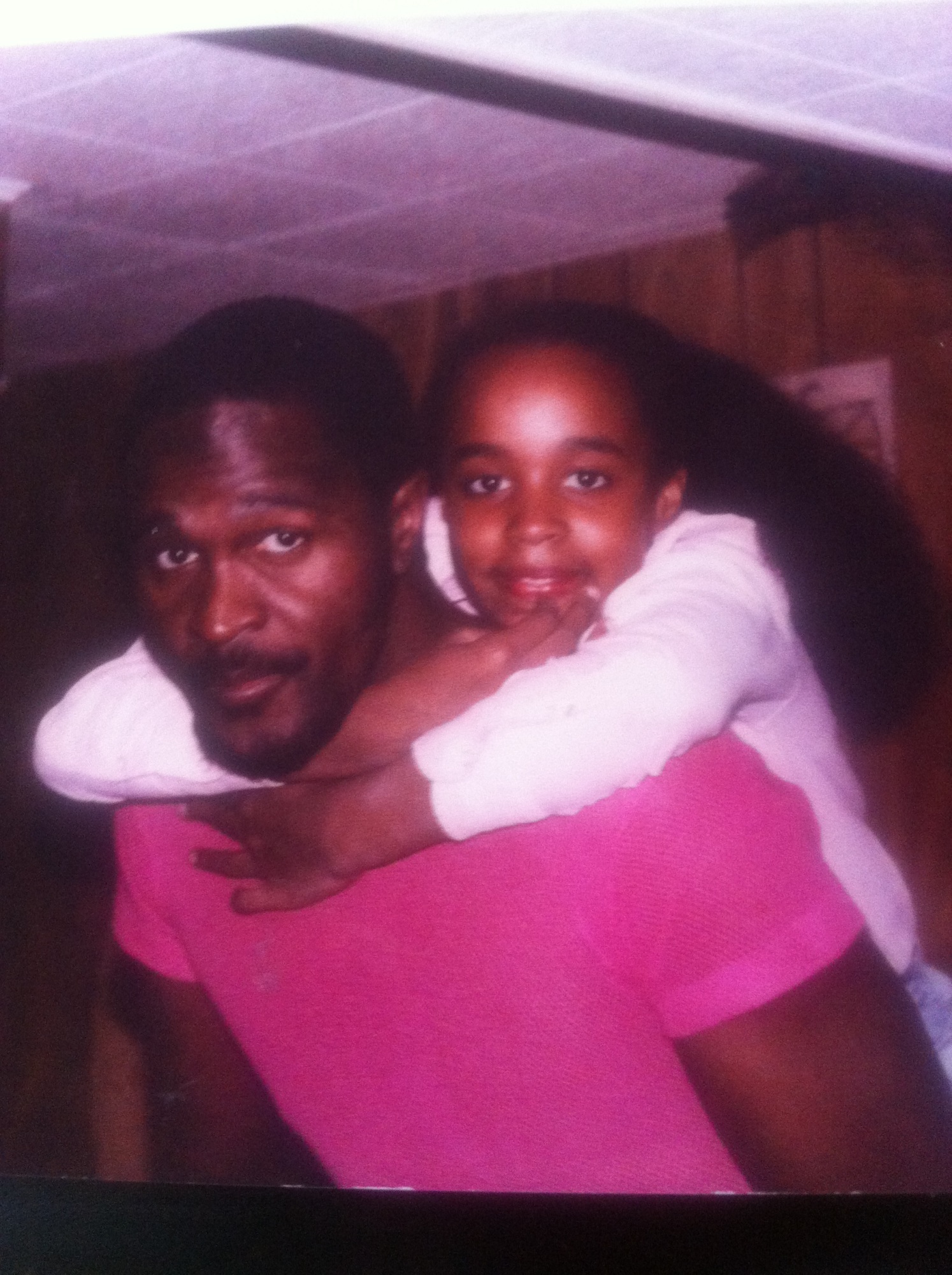 A man in a pink shirt gives a young girl a piggyback ride indoors. Both are looking at the camera and smiling.