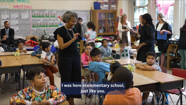 A teacher talks to elementary students seated at desks in a colorful classroom, while other adults and children are visible in the background. Caption reads: "I was here in elementary school, just like you.