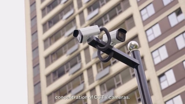 Two CCTV cameras mounted on a black pole in front of a tall apartment building; subtitle text reads, "concentration of CCTV cameras.