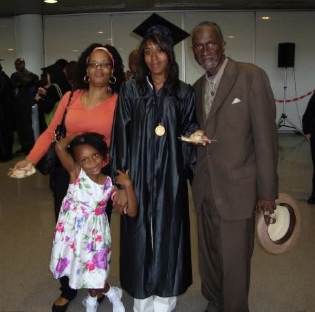 Three adults and a child pose indoors. The central woman wears a graduation cap and gown with a medal, holding a plate of food. The child in a floral dress poses playfully in front.