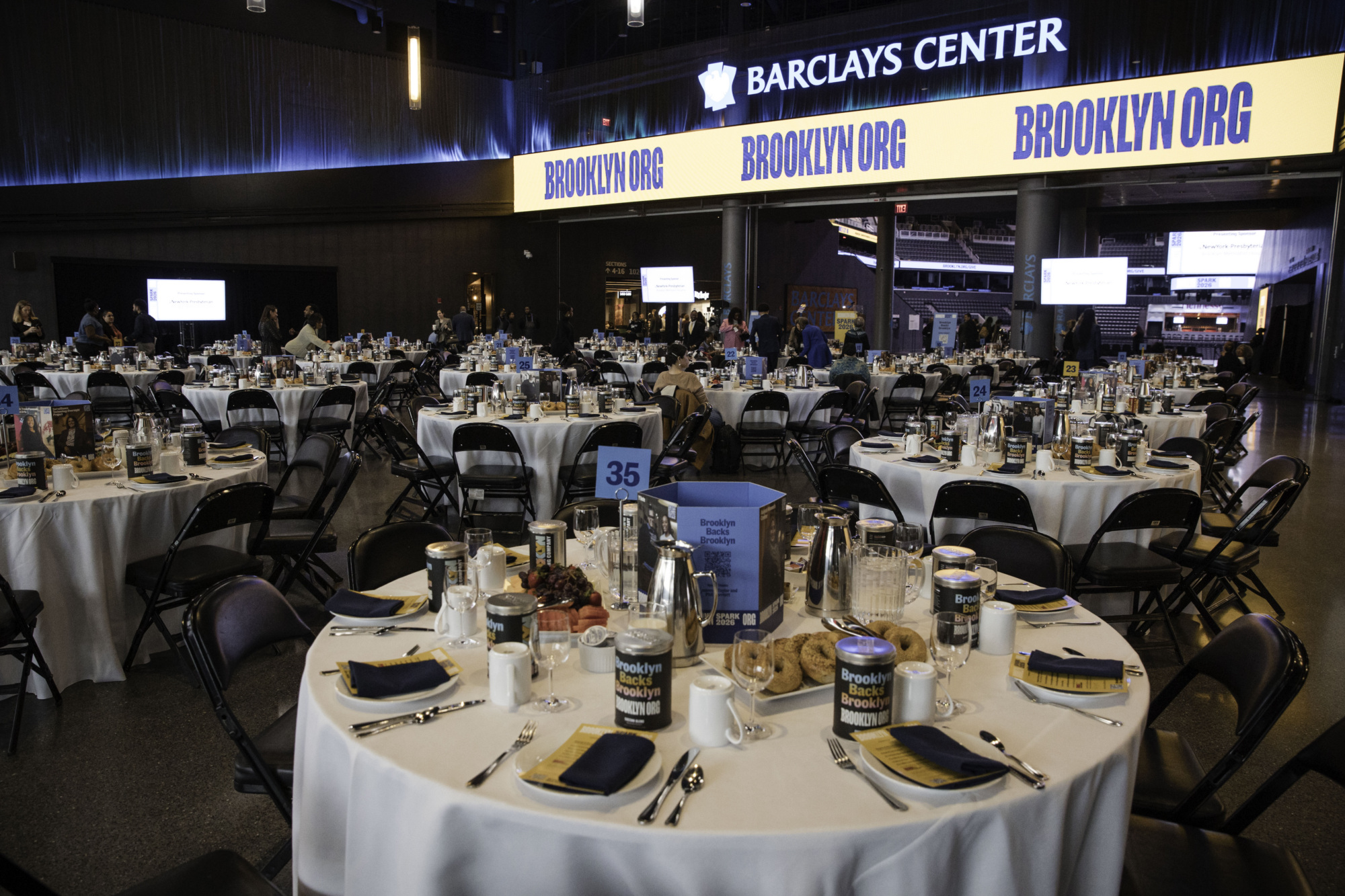 Round tables set for an event with place settings, programs, and food at Barclays Center; large screens and Brooklyn Org banners are visible in the background.