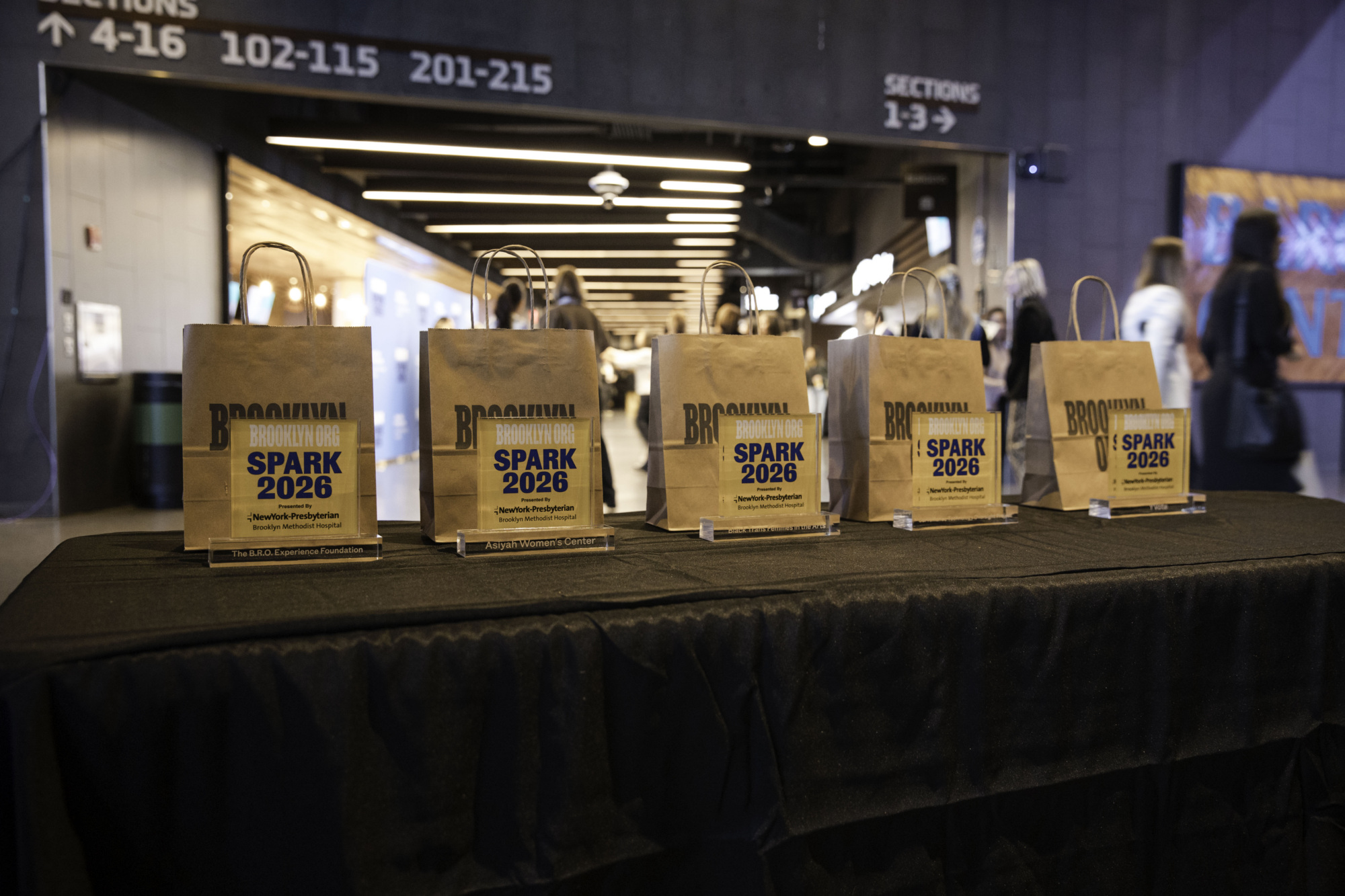 Five brown gift bags labeled "SPARK 2026" are displayed in a row on a black tablecloth in a public indoor area with people in the background.