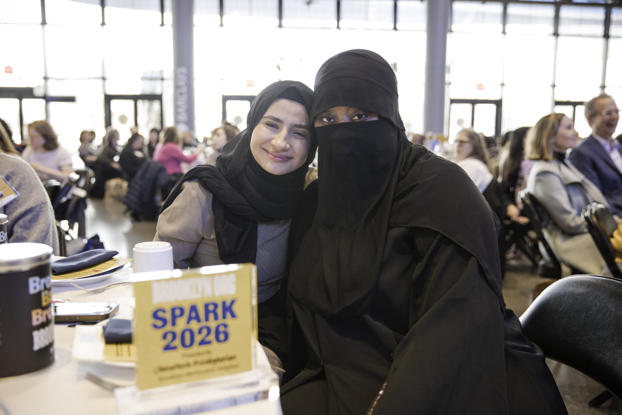 Two women sitting together at a conference table, one wearing a hijab and the other in a niqab, with event materials labeled "SPARK 2026" visible on the table.