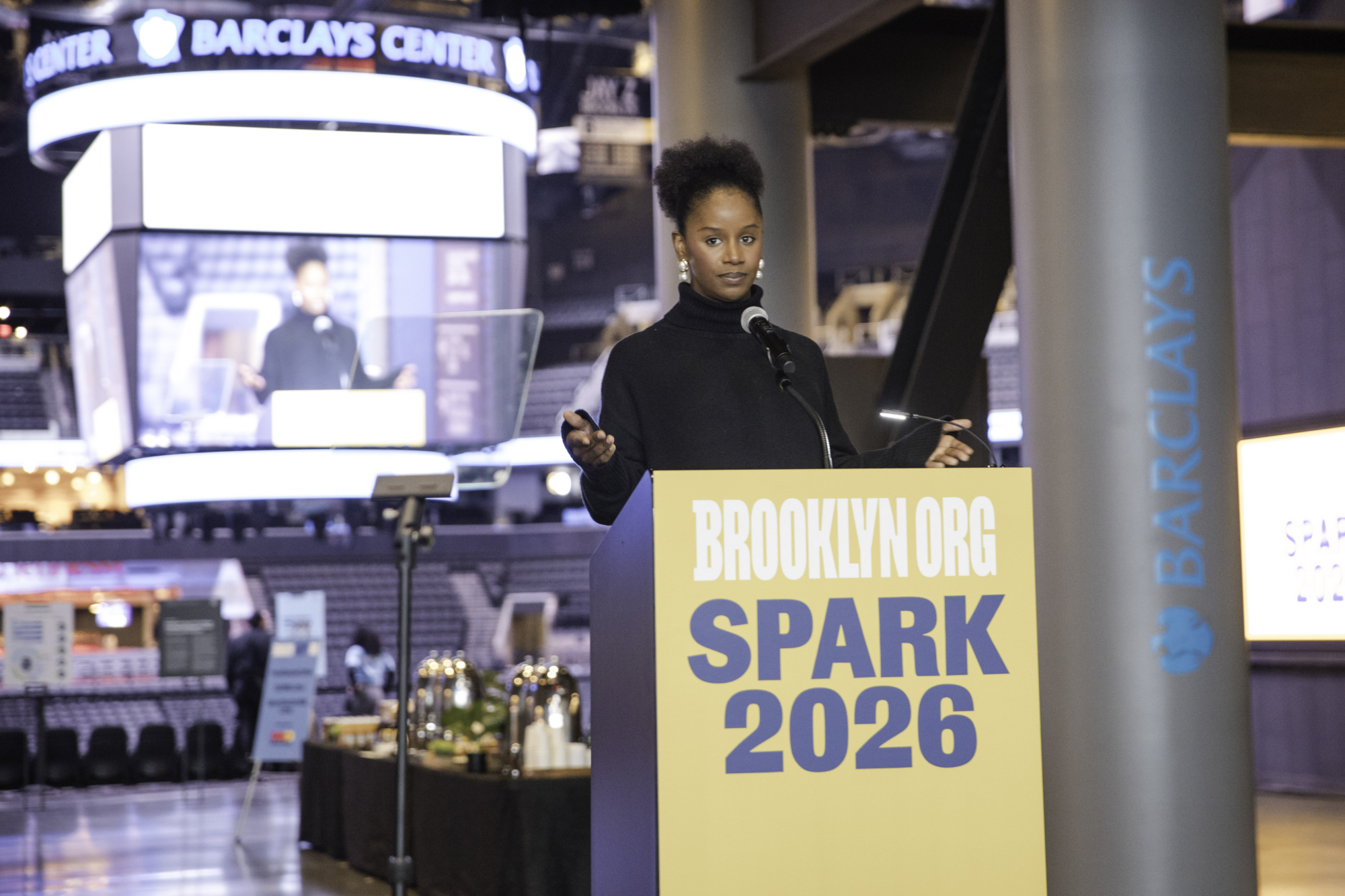 A person stands at a podium labeled "BROOKLYN.ORG SPARK 2026" speaking at an event inside the Barclays Center.