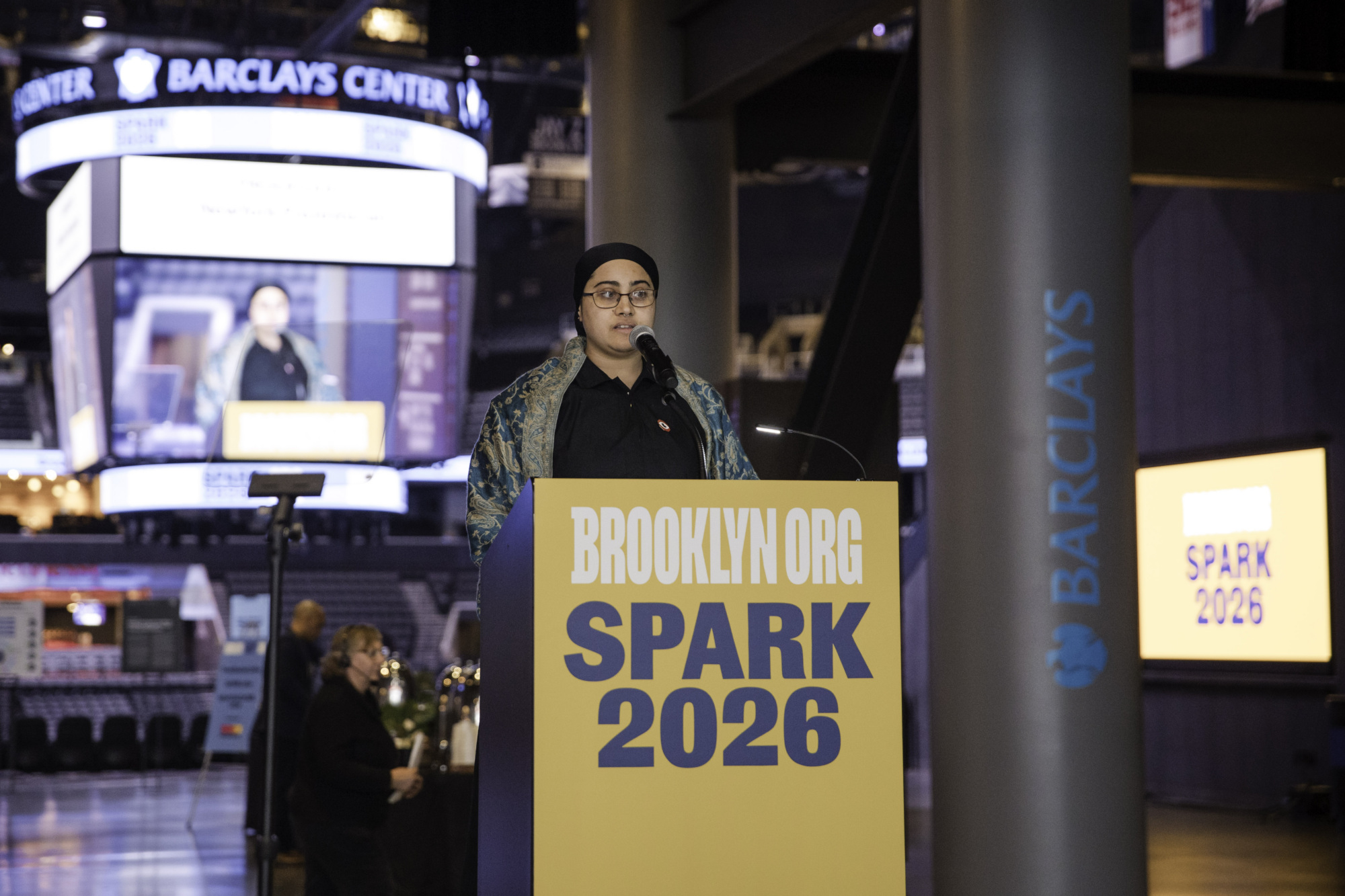 A person speaks at a podium labeled "Brooklyn.org Spark 2026" inside Barclays Center, with event branding visible in the background.