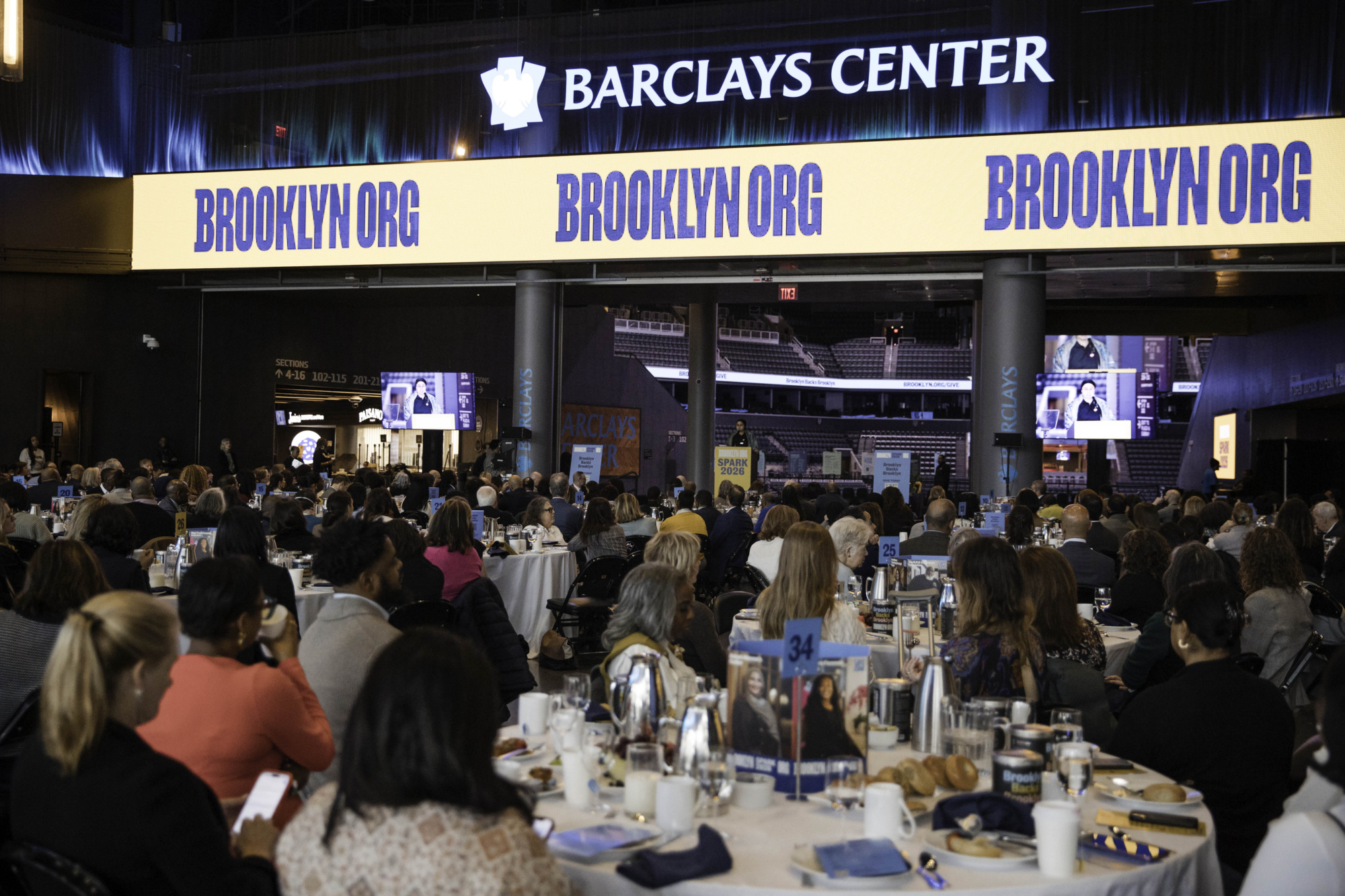 A large group of people sit at round tables during an indoor event at Barclays Center, with "BROOKLYN.ORG" displayed on screens above.
