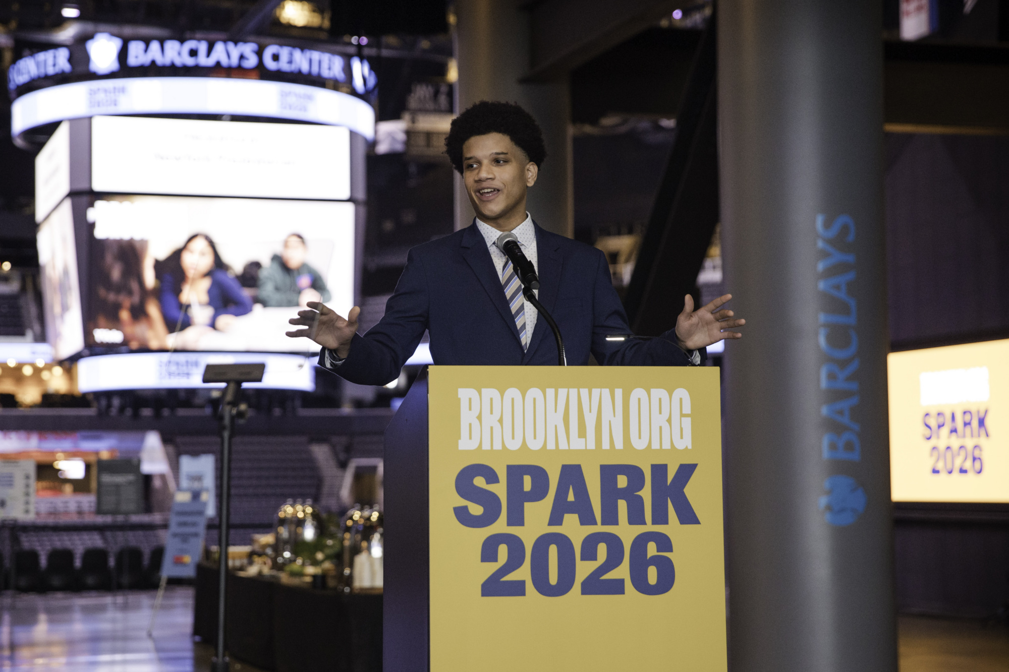 A man in a suit speaks at a podium labeled "Brooklyn.Org Spark 2026" inside Barclays Center, with event signage visible in the background.