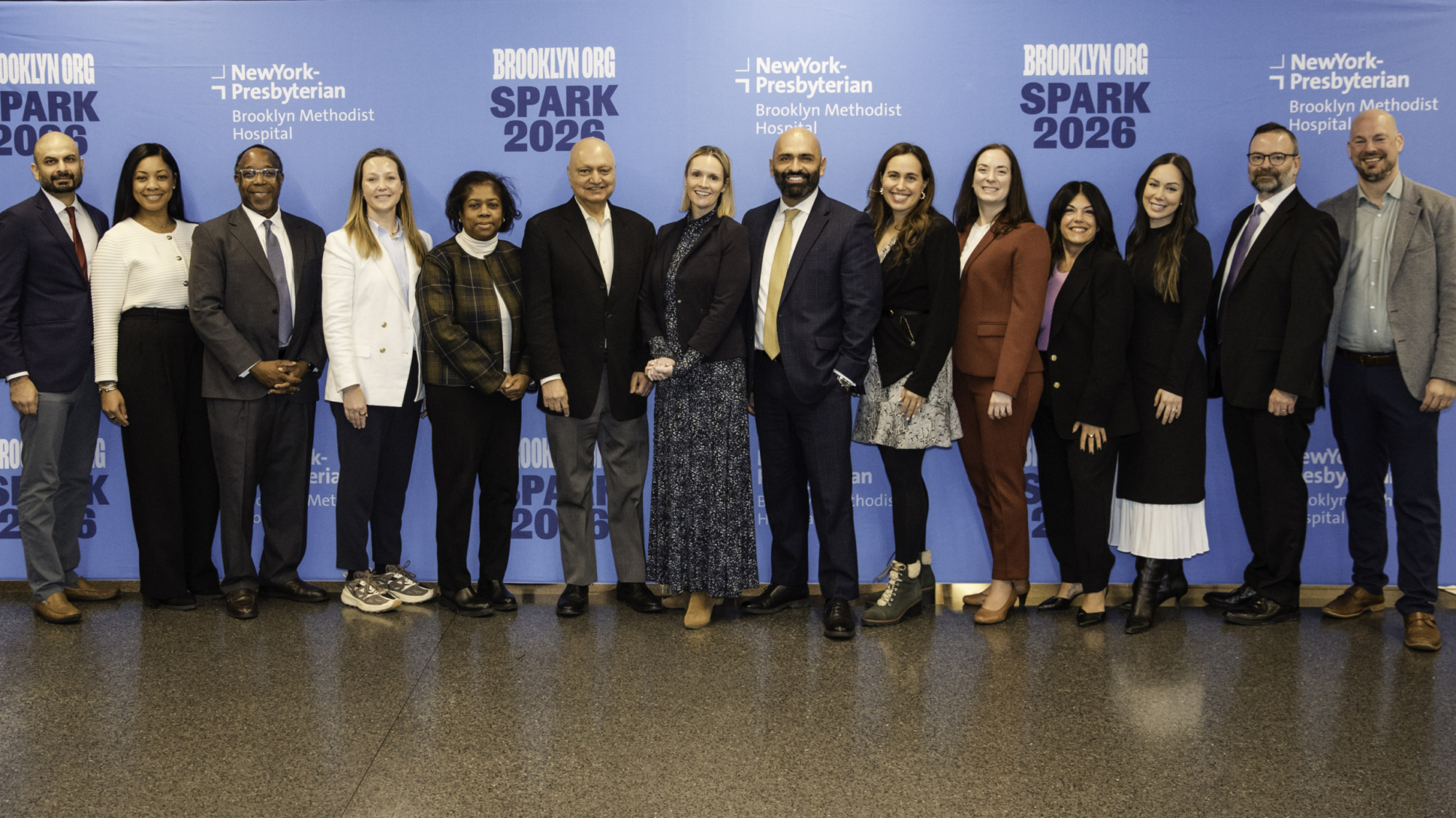 A group of thirteen people stand in front of a blue backdrop with "SPARK 2026" and "NewYork-Presbyterian Brooklyn Methodist Hospital" logos, posing for a group photo.