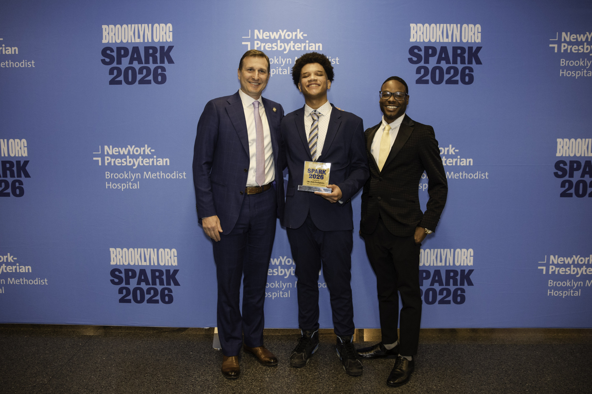 Three men in suits stand in front of a blue "Brooklyn Org SPARK 2026" step-and-repeat banner; the man in the middle holds a SPARK award plaque.