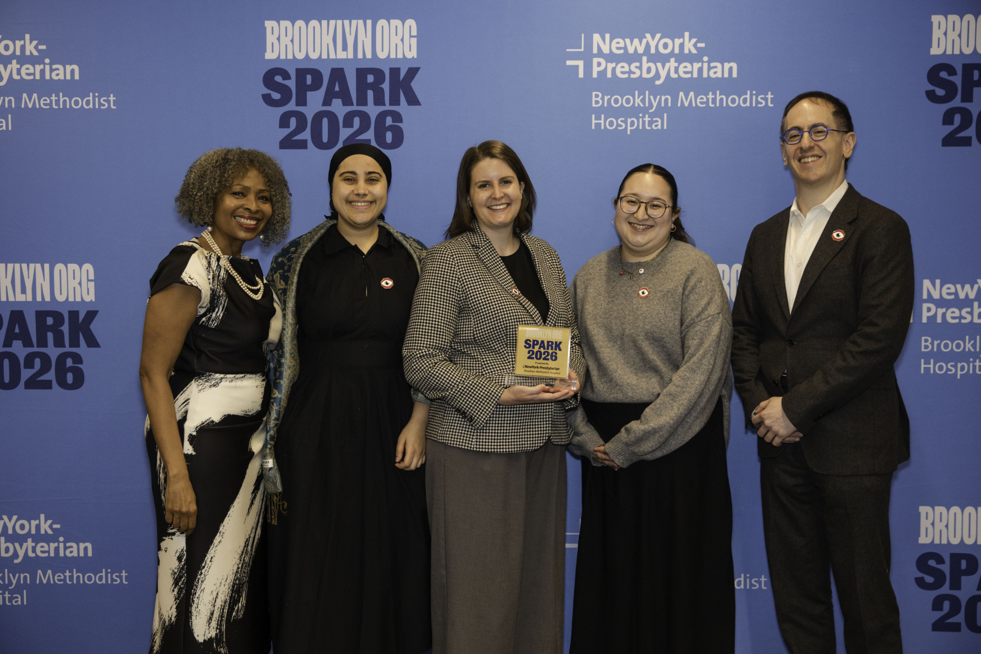 Five people stand in front of a blue Brooklyn Org SPARK 2026 and NewYork-Presbyterian backdrop, with one person holding a SPARK 2026 award.