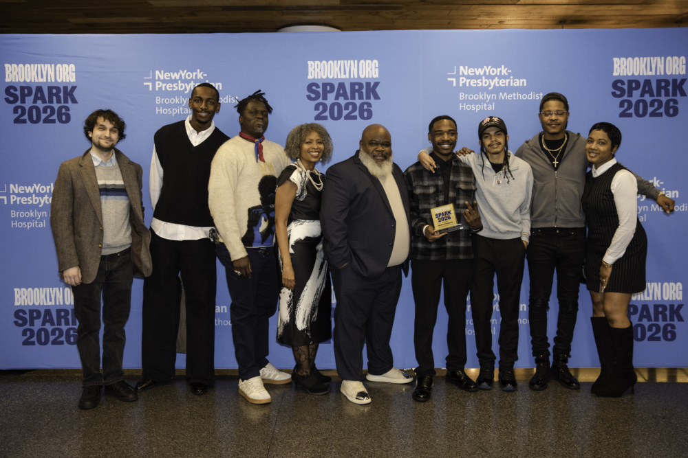 A group of eight people stands in front of a blue Brooklyn.org SPARK 2026 backdrop, with one person holding a small award plaque.