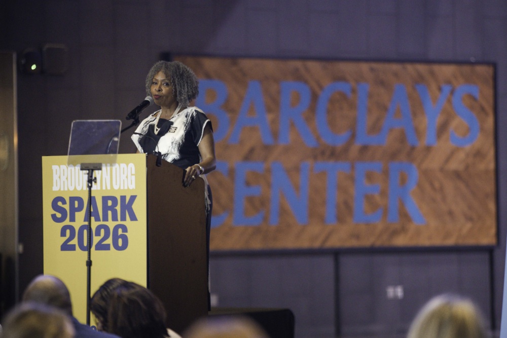 A woman stands at a podium labeled "SPARK 2026" speaking to an audience, with a large "Barclays Center" sign visible in the background.