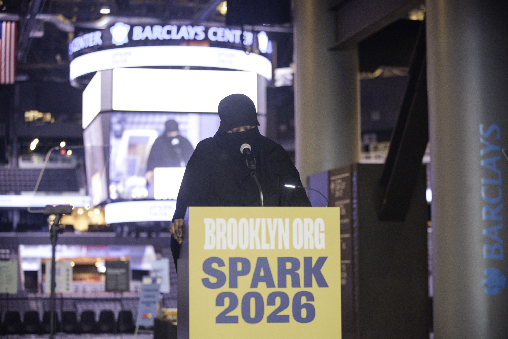 A person in black attire stands at a podium labeled "BROOKLYN.ORG SPARK 2026" inside the Barclays Center arena.