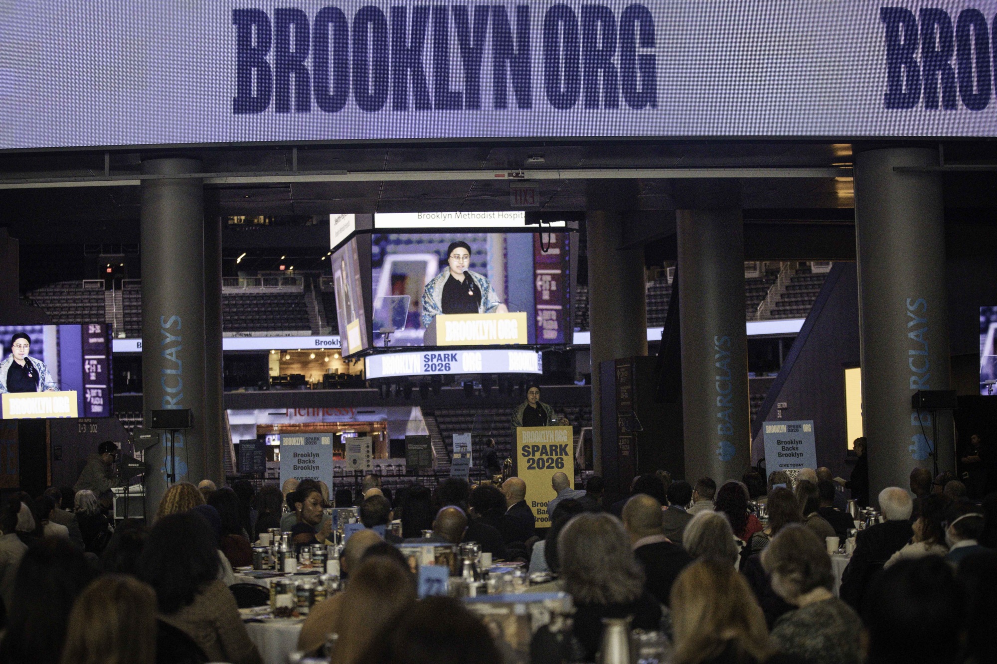 A speaker stands at a podium labeled "SPARK 2026" addressing a seated audience at an indoor event under a large "BROOKLYN.ORG" sign.