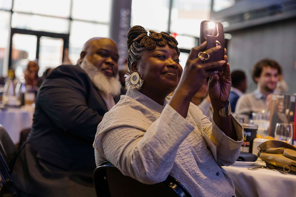 A woman takes a photo with her phone while seated at a table during an indoor event, with other attendees visible in the background.