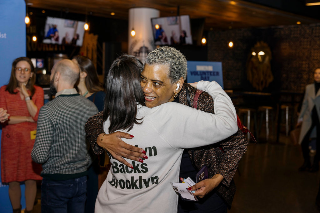Two people hug at an indoor event; one wears a sweatshirt reading "Brown Backs Brooklyn" while others converse in the background.