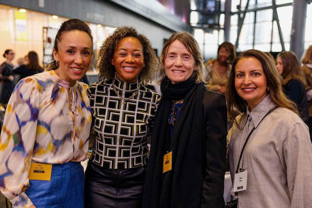Four women stand together indoors at an event, smiling at the camera and wearing name badges. Other people are visible in the background.