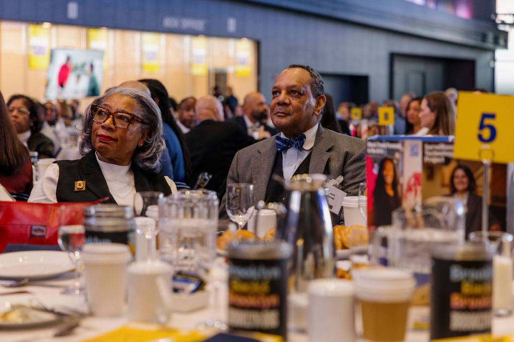 Two people sit at a banquet table during a formal event, surrounded by food, drinks, and event materials, with other attendees visible in the background.