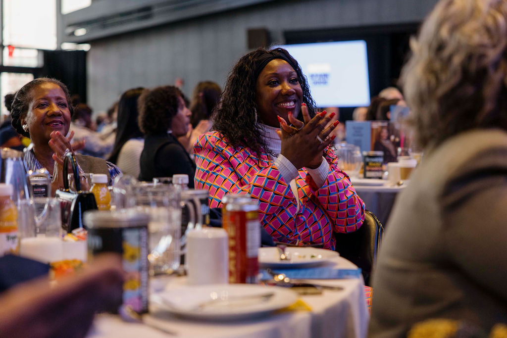 A group of people sit at round tables during an indoor event, with one woman in a patterned jacket clapping and smiling.