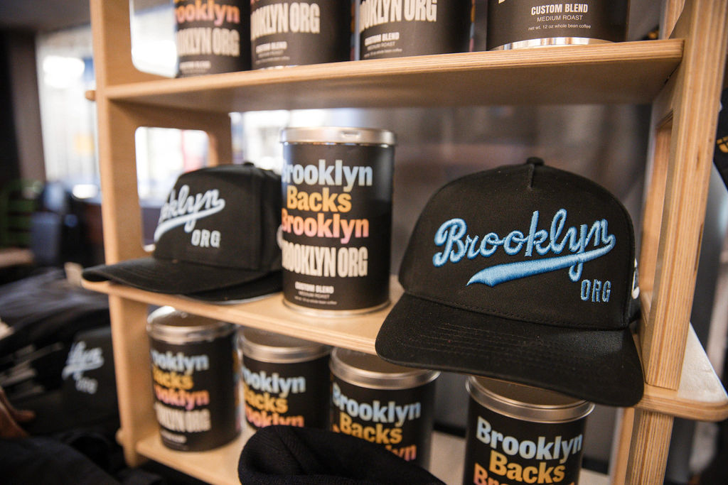 Display shelf with black "Brooklyn Org" hats and cylindrical containers labeled "Brooklyn Backs Brooklyn" in a store setting.