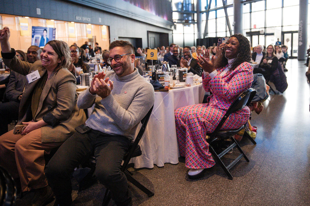 A group of people seated at round tables indoors clap and smile during an event or presentation.