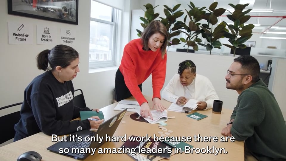 Four people sit around a table in an office, discussing papers. A woman in red stands and gestures toward documents. Subtitles read: “But it’s only hard work because there are so many amazing leaders in Brooklyn.”.