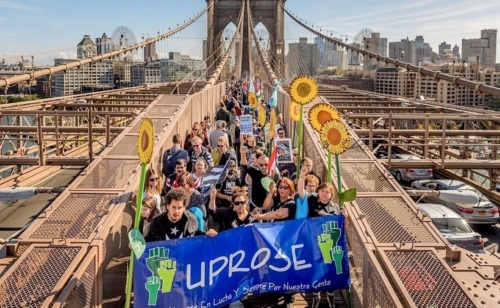 A group of people march across the Brooklyn Bridge holding sunflowers and a blue UPROSE banner, with city buildings in the background.