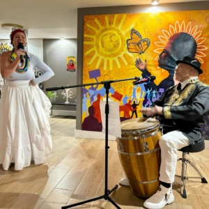 A woman sings into a microphone while a seated man plays a conga drum. Colorful artwork featuring sun and figures is displayed on the wall behind them.