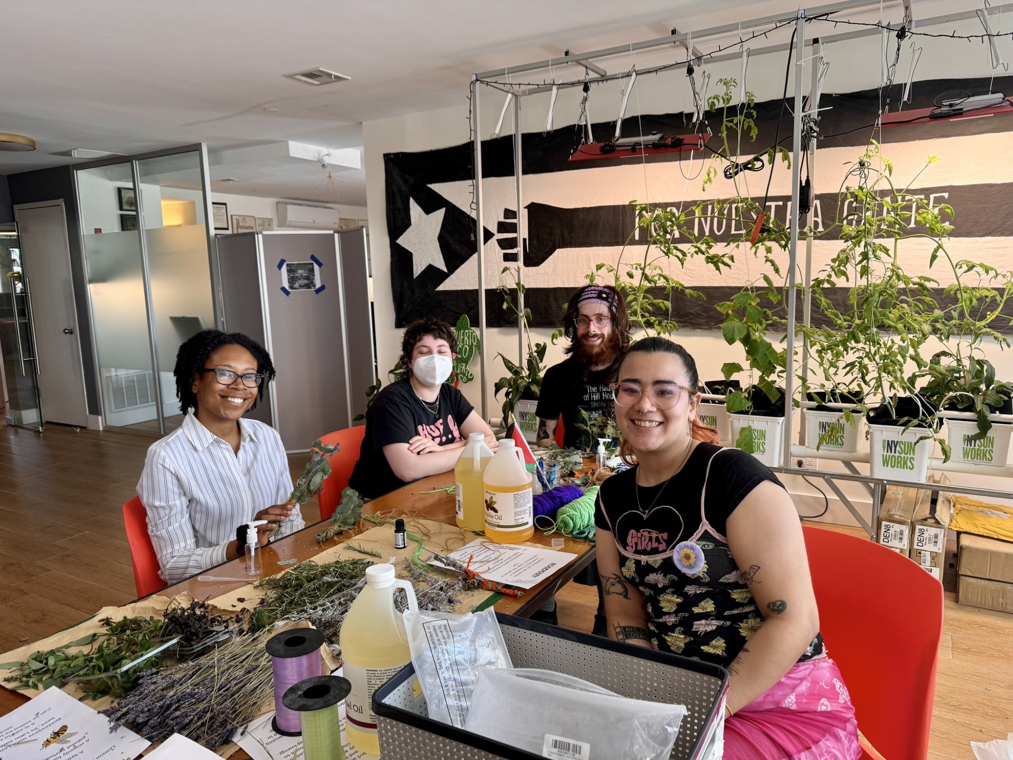 Four people sit at a table with plants, bottles, and craft supplies, smiling at the camera in a room with a large black and white flag and potted plants in the background.