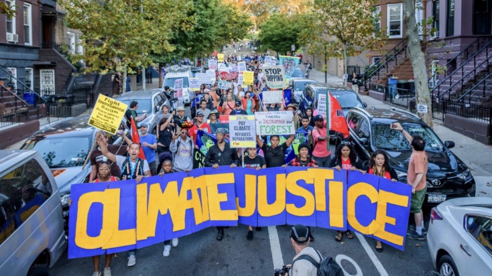 A large group of people march down a city street holding signs, including a prominent banner reading “Climate Justice.”.