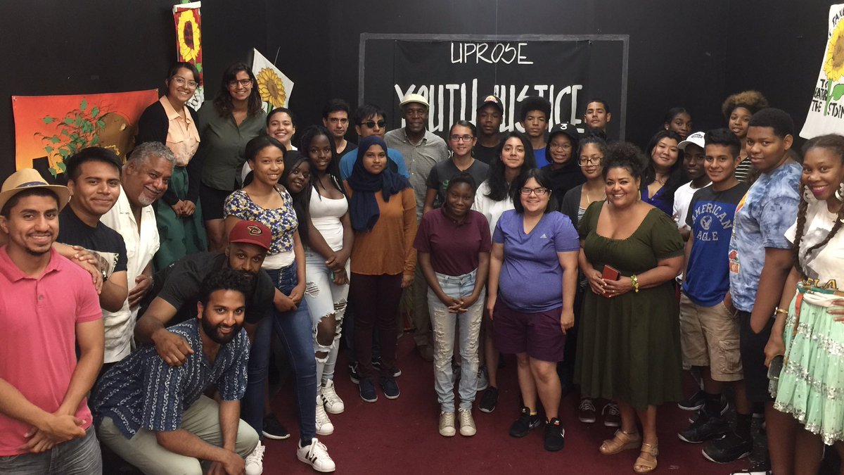 A diverse group of people poses together in a room with colorful art and a sign that reads “UPROSE Youth Justice” on the wall behind them.