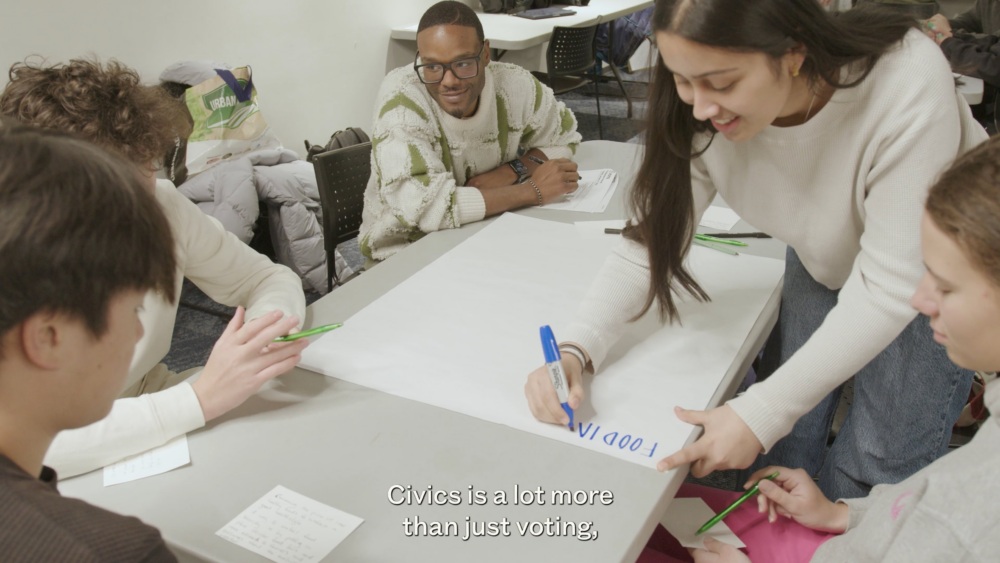 Four people sit around a table; one woman is writing "FOOD" on a large sheet of paper. Subtitles read: "Civics is a lot more than just voting.