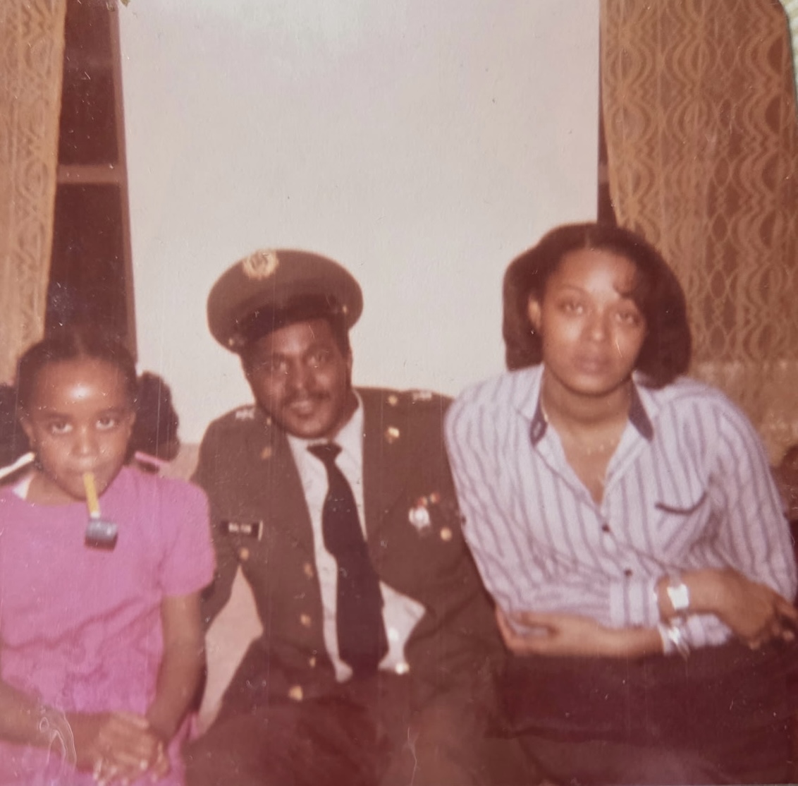 A young girl with a toy pipe, a man in a military uniform, and a woman in a striped shirt sit together indoors in front of curtained windows.