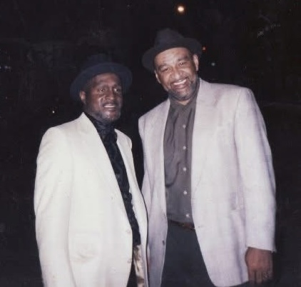 Two men stand together at night, both wearing light-colored jackets, dark shirts, and hats, facing the camera and smiling.