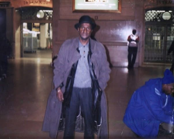 An older man wearing a hat and trench coat stands indoors, with another person in blue bending down nearby; background includes lighted signage and closed shop gates.