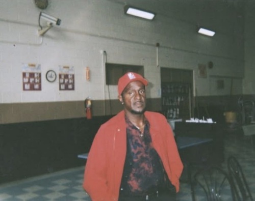 A man wearing a red jacket and matching cap stands indoors in a room with tiled floors, wall clocks, and various furniture items in the background.