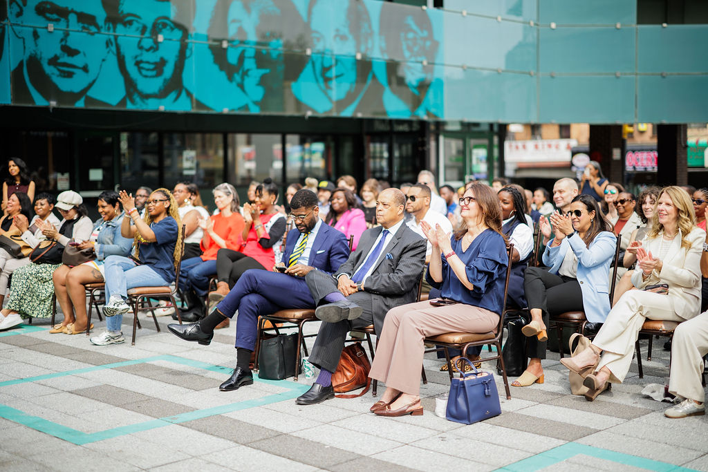 Audience seated outdoors, attentively watching and clapping during an event in front of a modern building with large mural portraits.