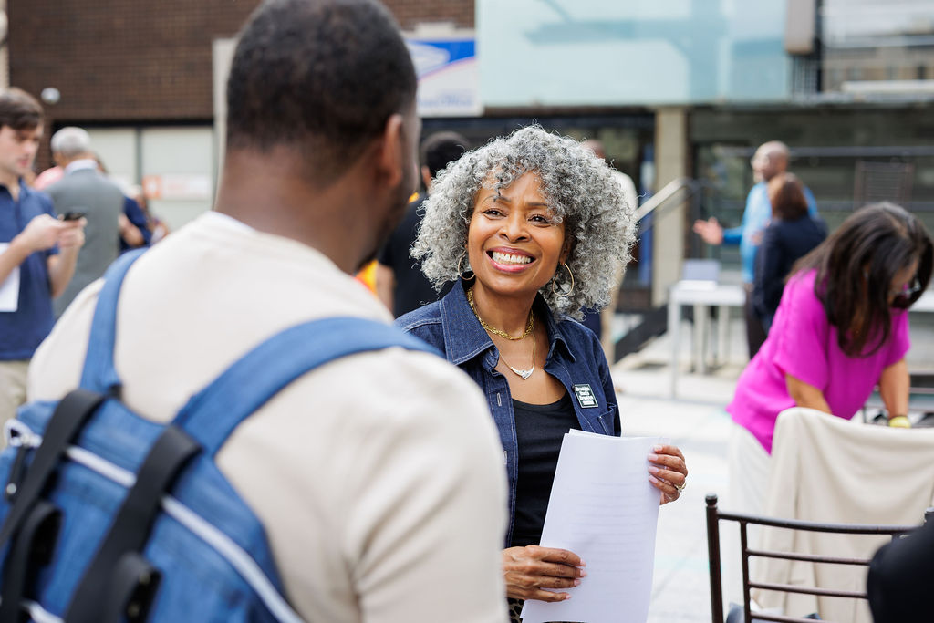 A woman with curly gray hair smiles while holding papers and talking to a man at an outdoor event with other people in the background.