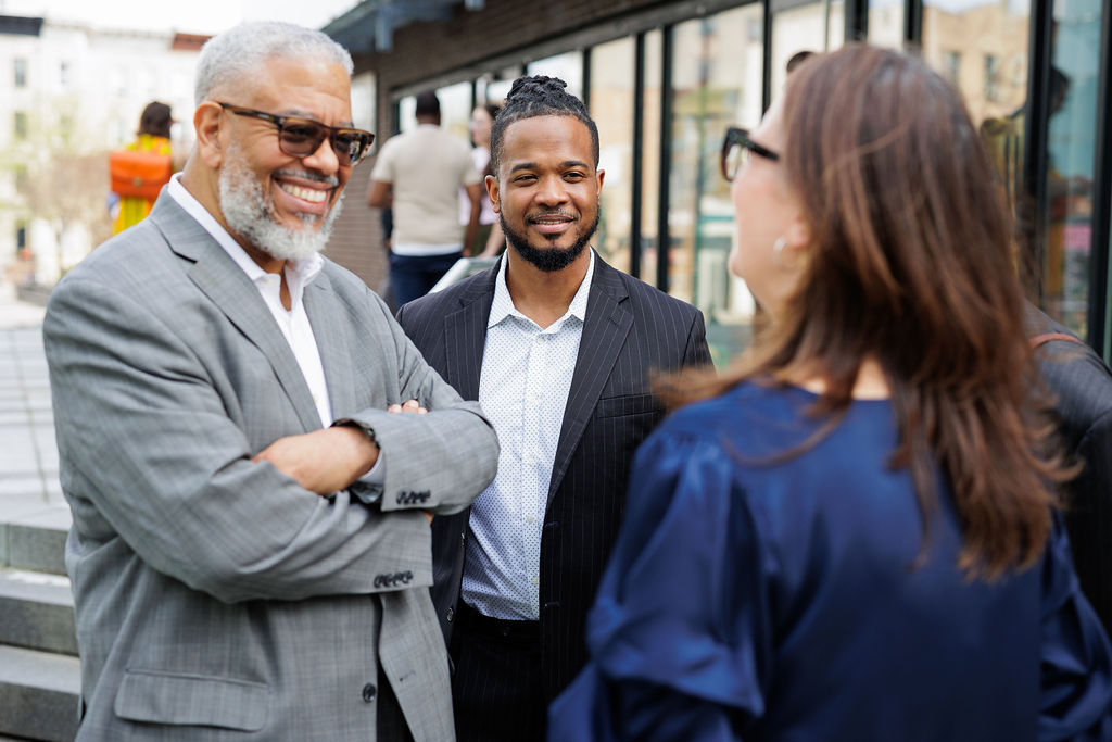 Three professionally dressed people stand outside, engaged in conversation. Two men face a woman with their backs to a modern building.