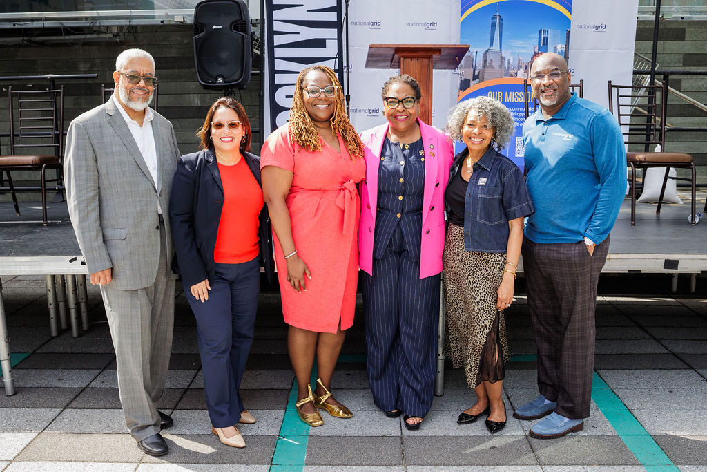Six adults stand side by side outdoors, smiling at the camera in front of a stage with chairs and city-themed banners.