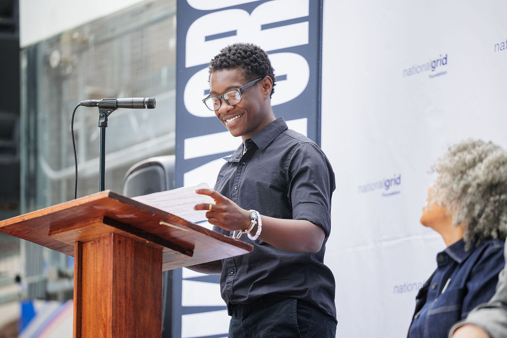 A person stands at a wooden podium speaking into a microphone, while another person sits nearby. A "National Grid" banner is visible in the background.