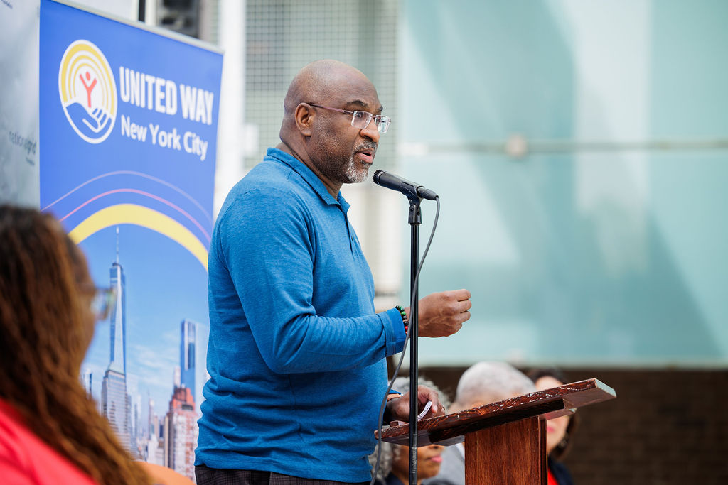 A man in a blue shirt speaks at a podium with a microphone, with a United Way New York City banner in the background.