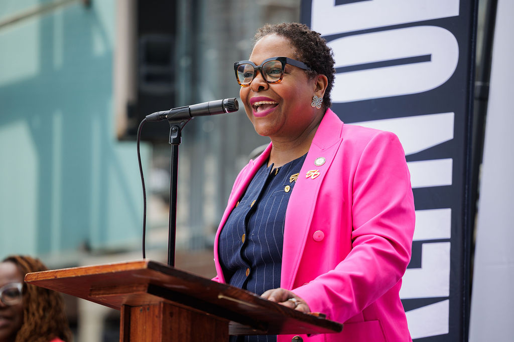 A woman wearing a bright pink blazer and glasses speaks at a podium with a microphone, standing in front of a large vertical sign.