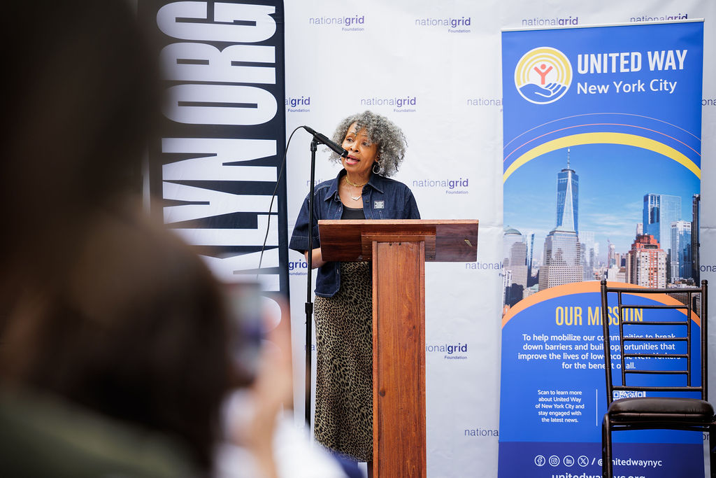 A woman speaks at a podium between a United Way New York City banner and a large sign, with a national grid backdrop behind her.
