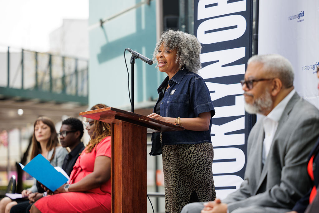 A woman speaks at a podium during an outdoor event, with several people seated behind her and a "Brooklyn" banner in the background.